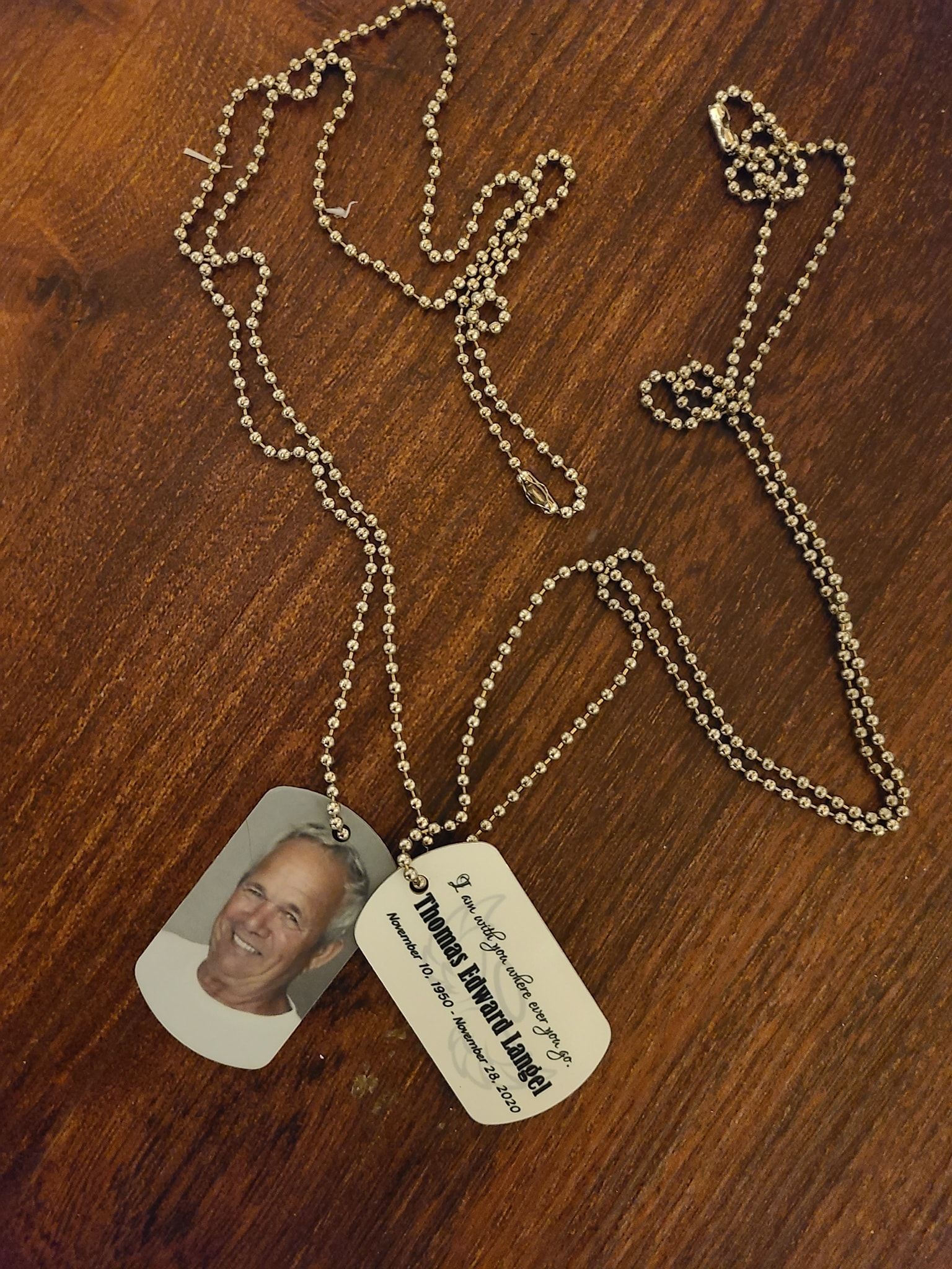 Two dog tags on chains with a man's photo on one and text on the other. They rest on a wood surface.