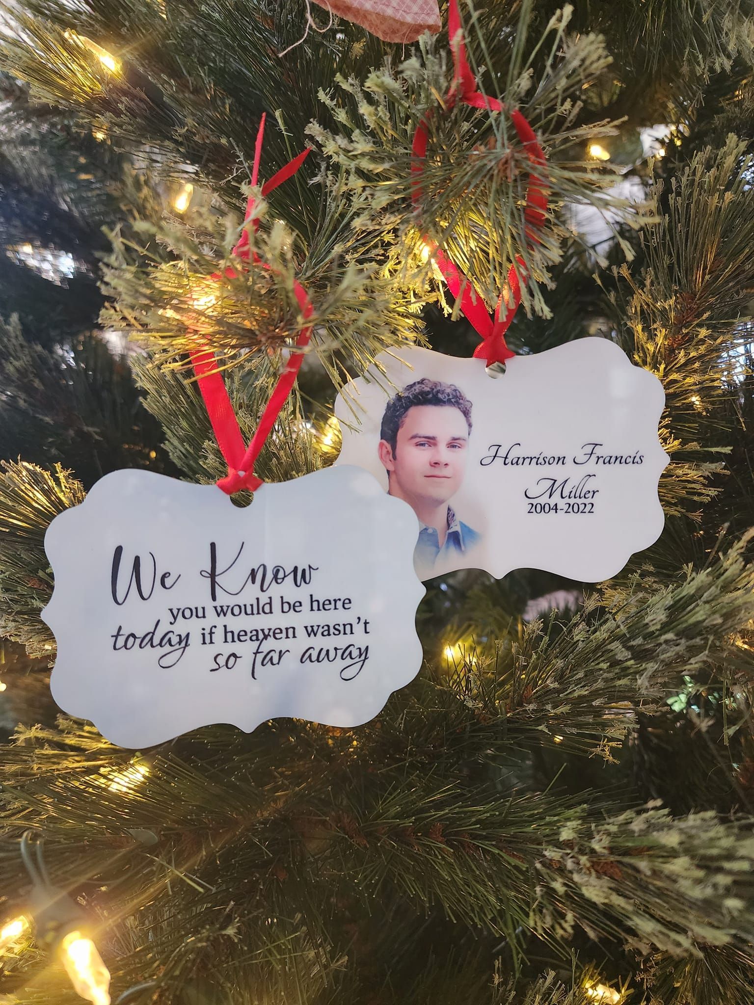 Two memorial ornaments on a Christmas tree. One has a photo and name, the other has text. Both hang with red ribbon.