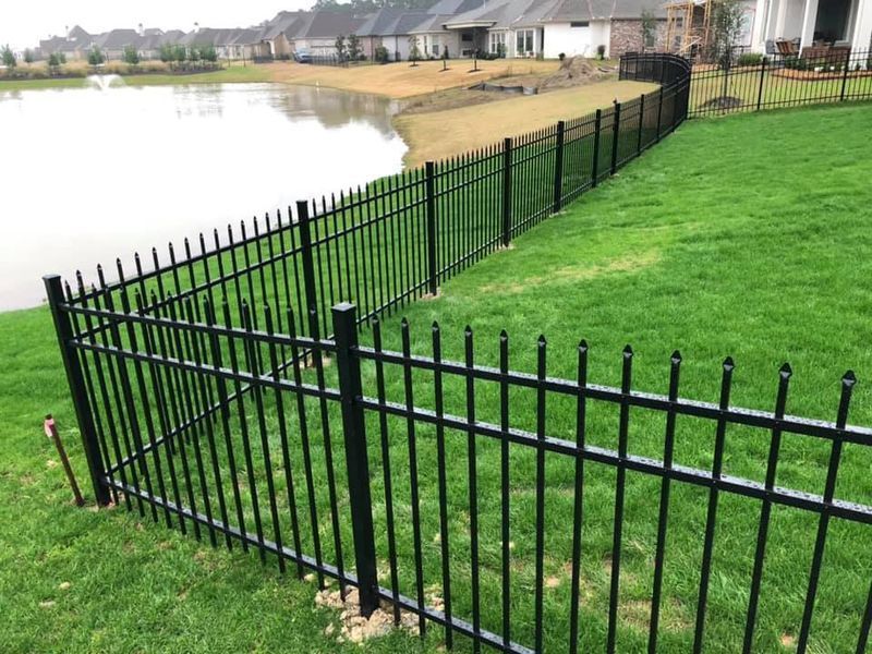 A black metal fence surrounds a lush green lawn next to a lake.