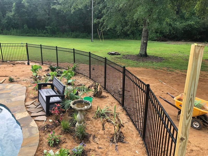 A black fence surrounds a garden with a bench and a bird bath.