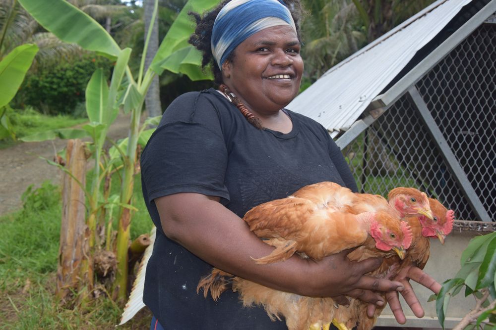 Une femme tient un poulet dans ses mains devant un poulailler.