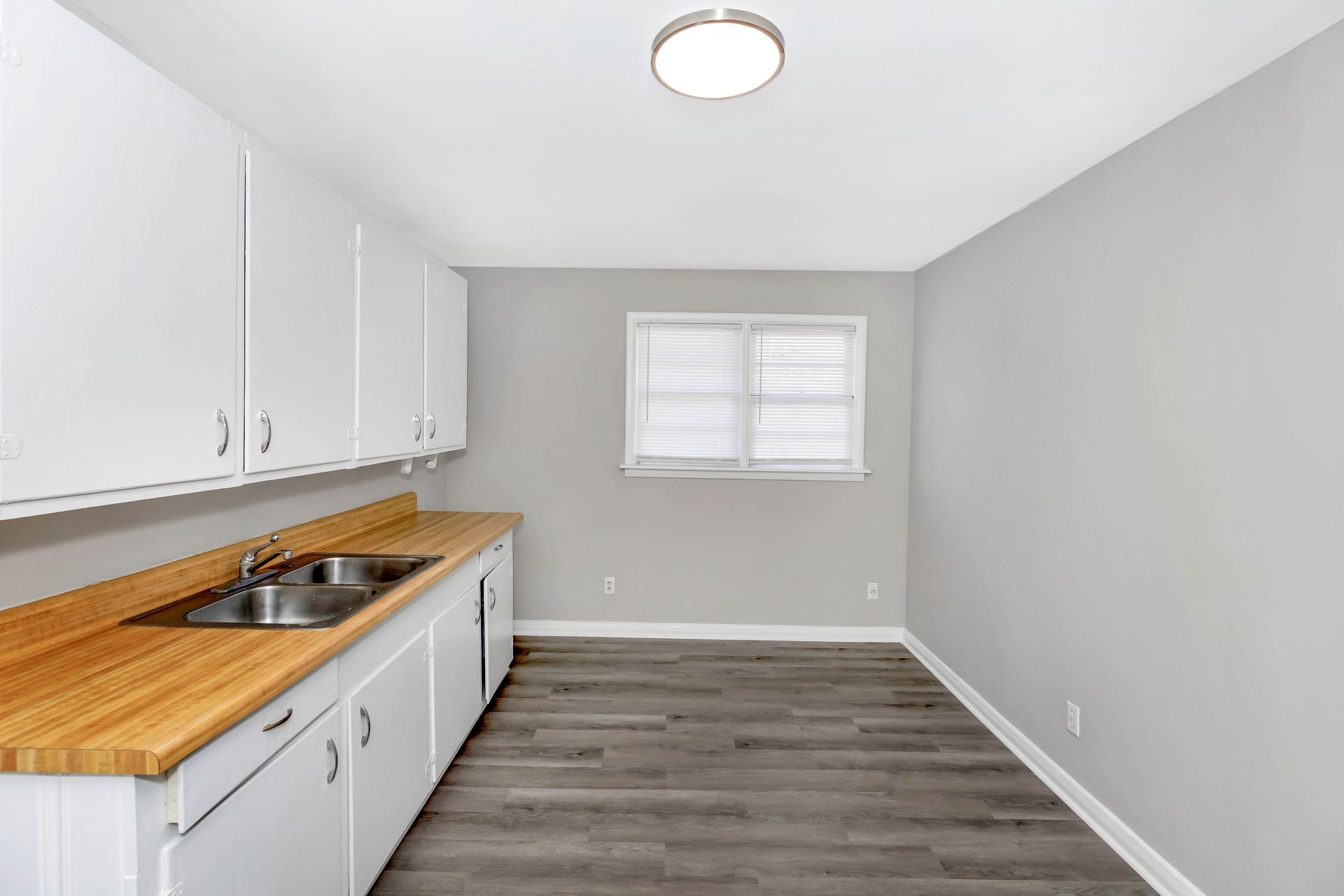 An empty kitchen with white cabinets , a sink , and a window.