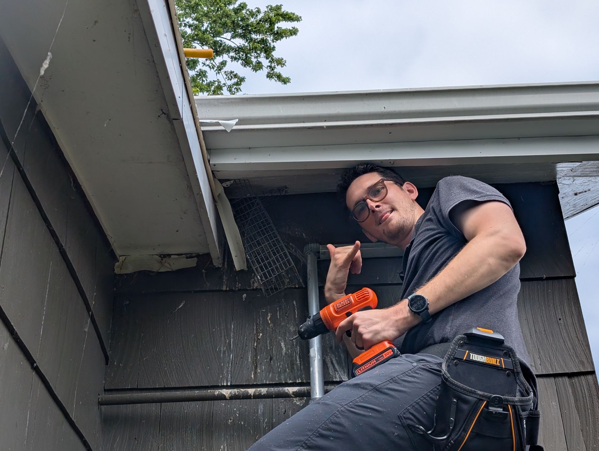 Man on ladder drills into a house's siding near a gutter; he points a thumb up and smiles, holding an orange power drill.