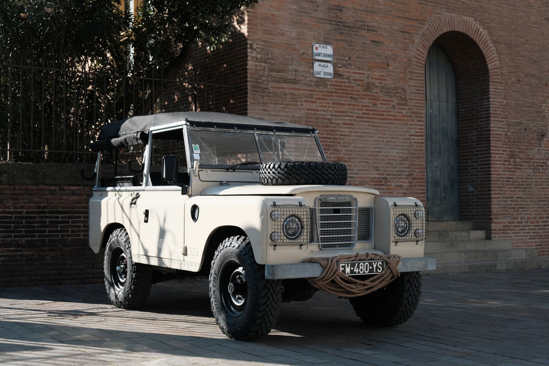 Land Rover beige garé sur une rue pavée, à côté d'un immeuble en briques. Roue de secours sur le capot, pneus noirs.