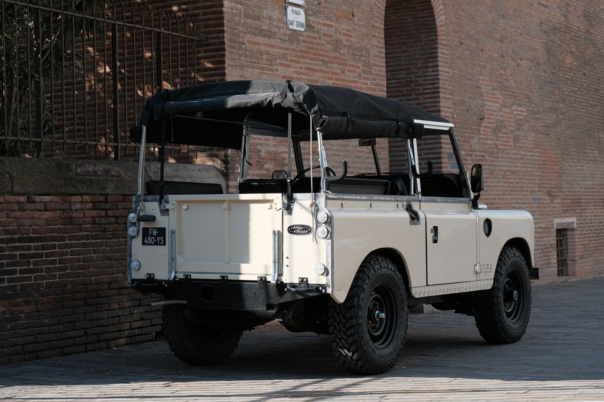 Land Rover Defender beige avec capote noire, garé à côté d'un mur de briques.