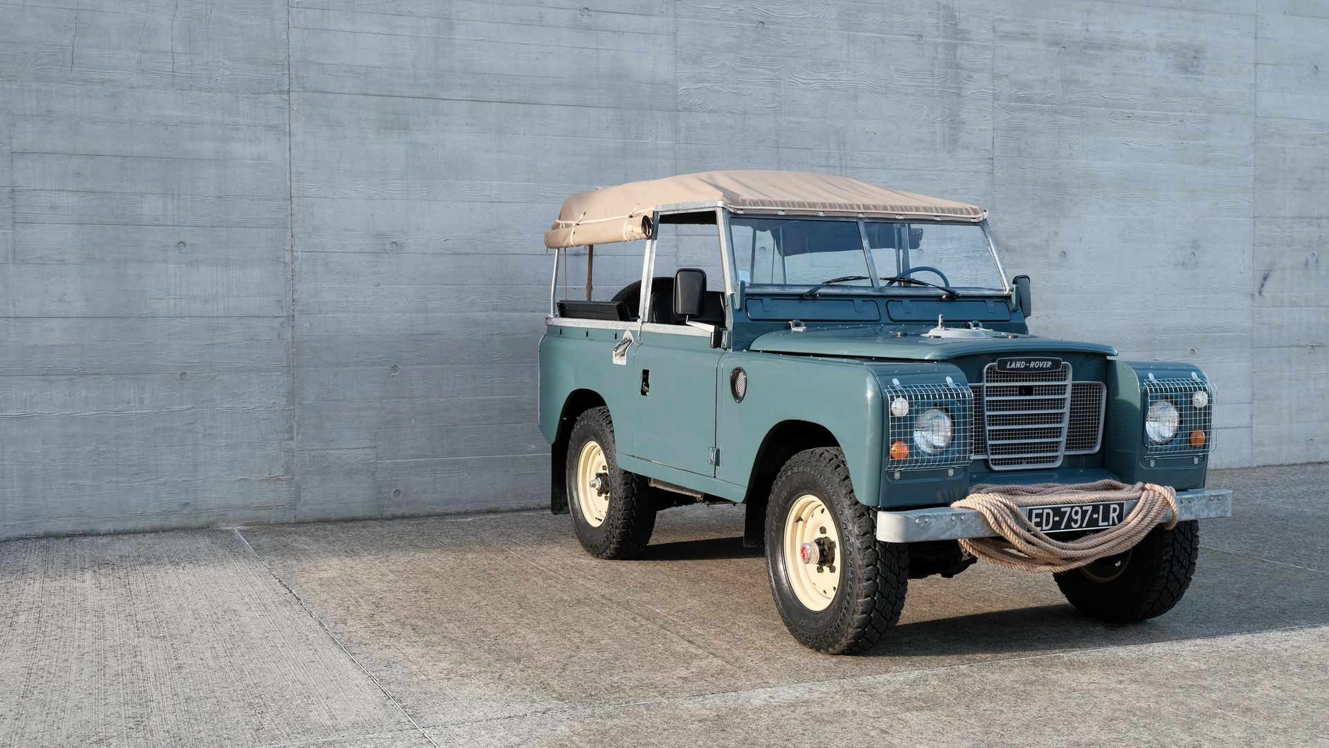 Land Rover bleu, toit beige, garé sur du béton, à côté d'un mur gris.