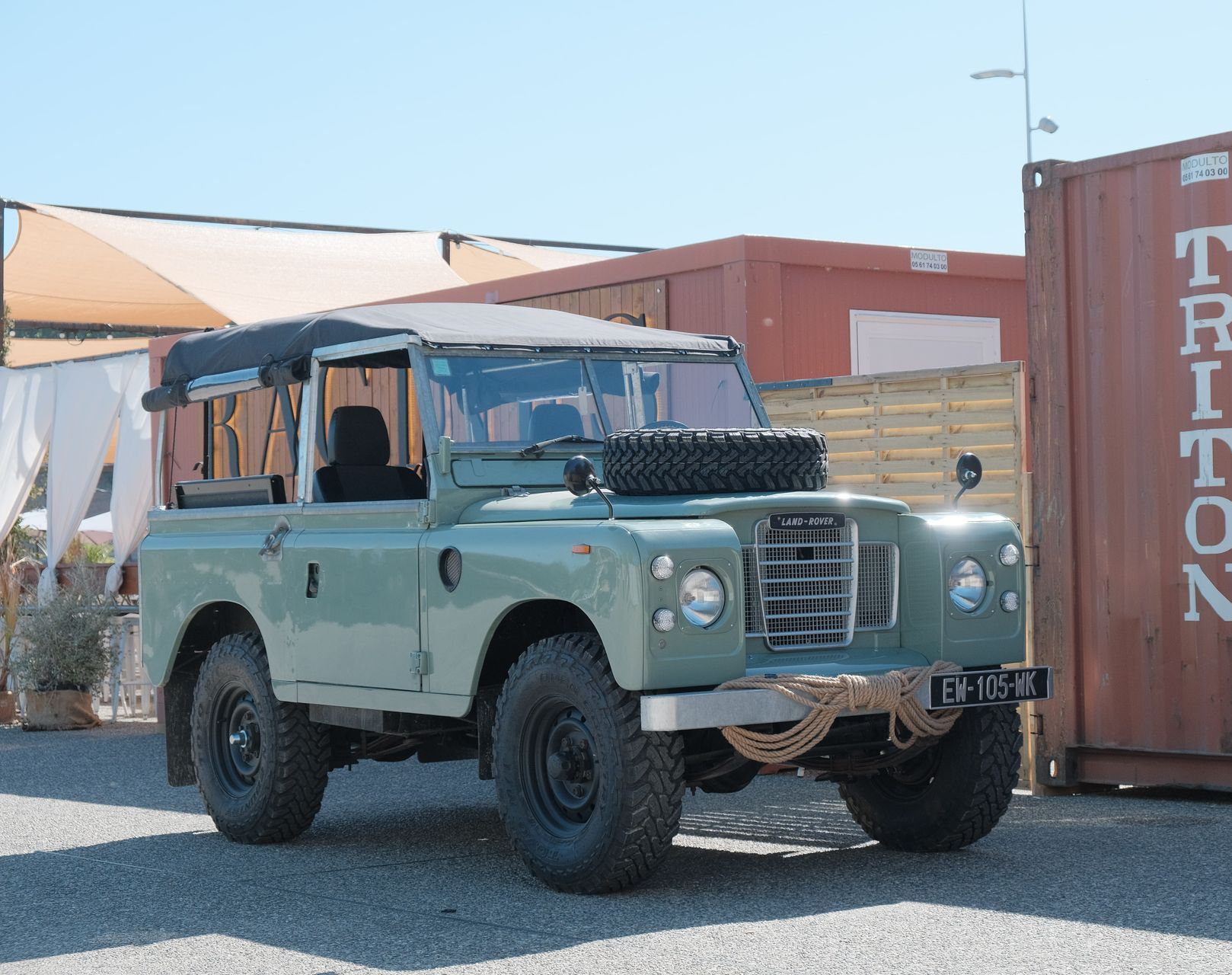 Land Rover vert vintage garé devant des conteneurs d'expédition par une journée ensoleillée.