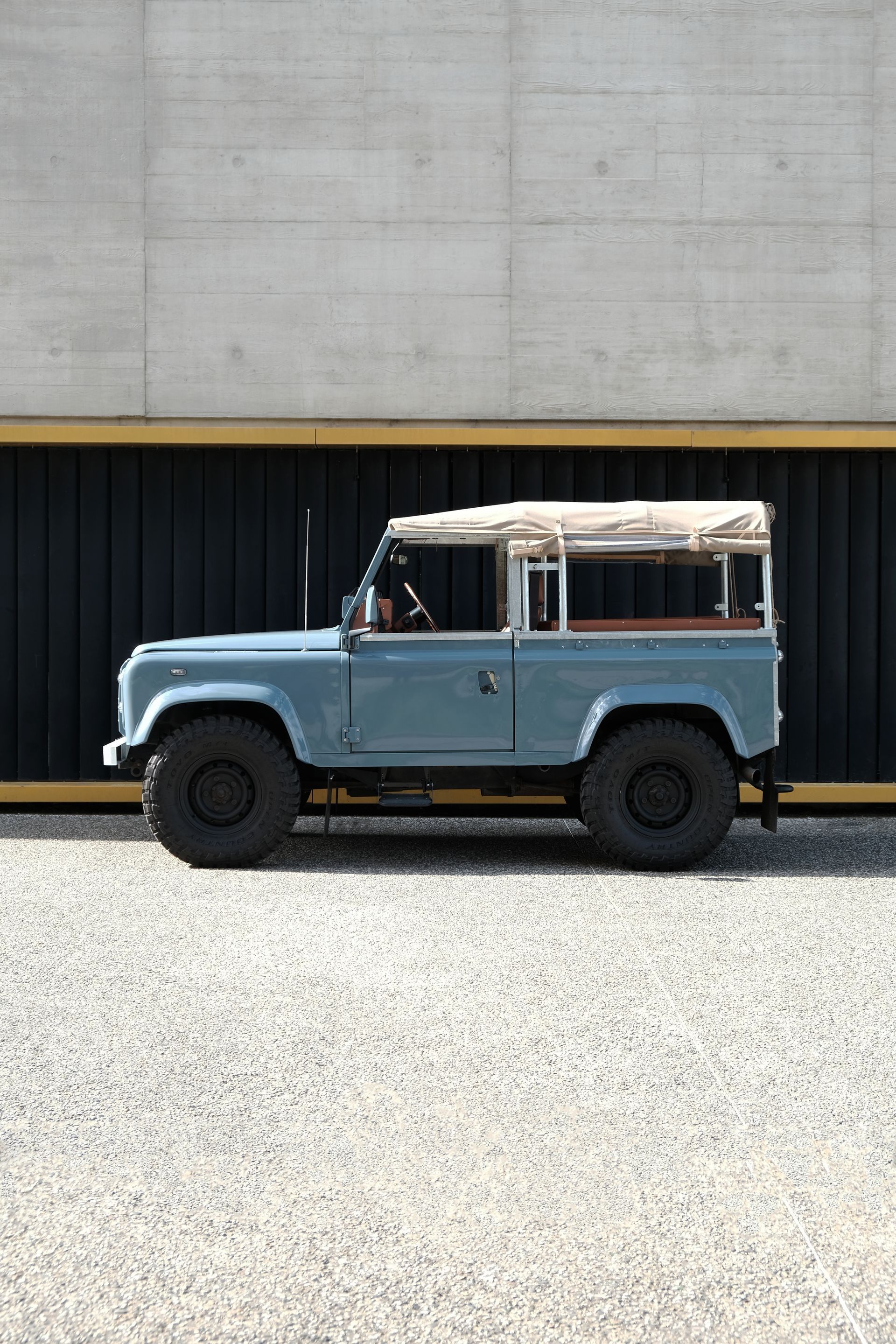 Land Rover Defender bleu clair avec un toit beige, garé sur du gravier.