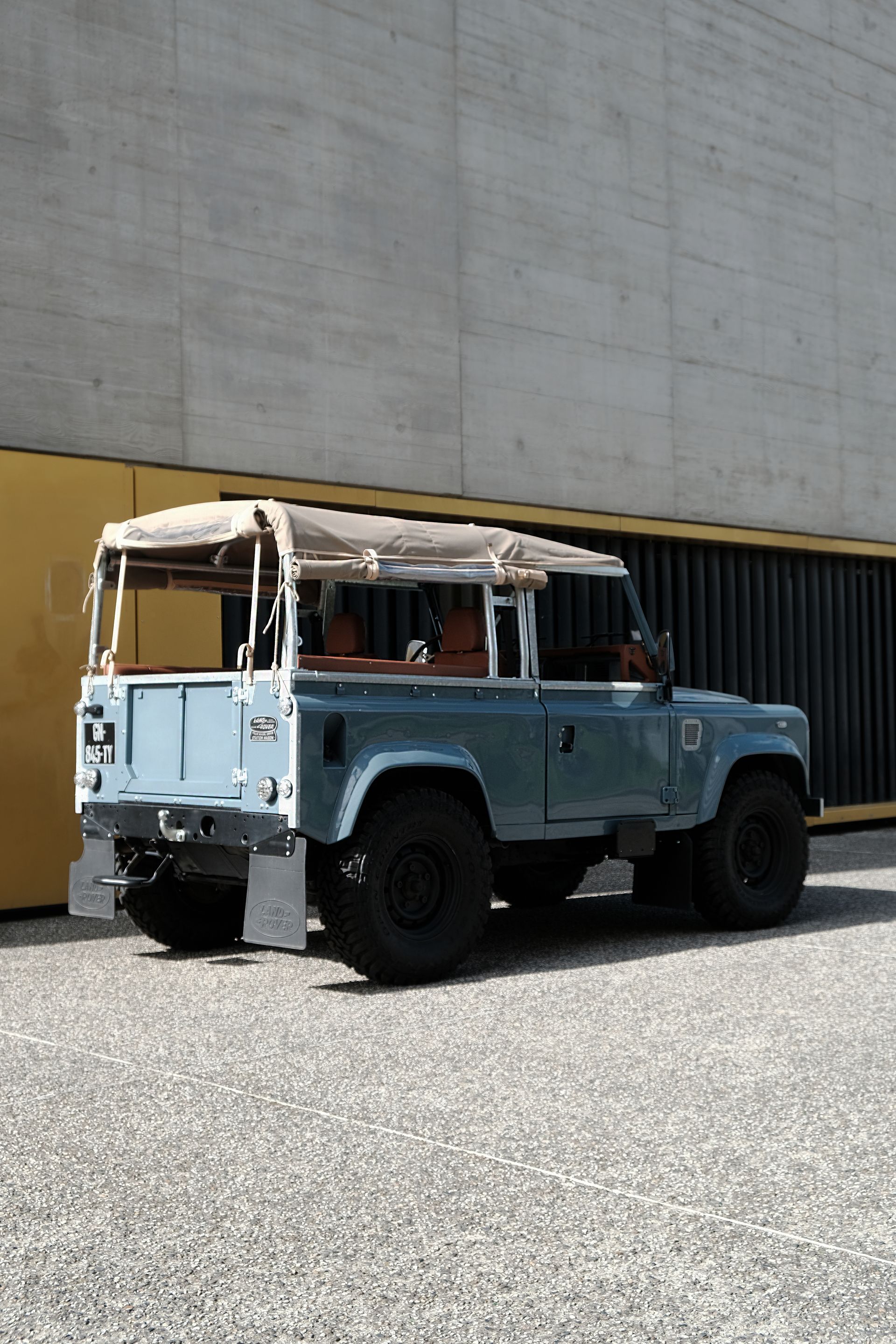 Land Rover Defender bleu vintage avec toit beige garé devant des murs en béton et jaunes.
