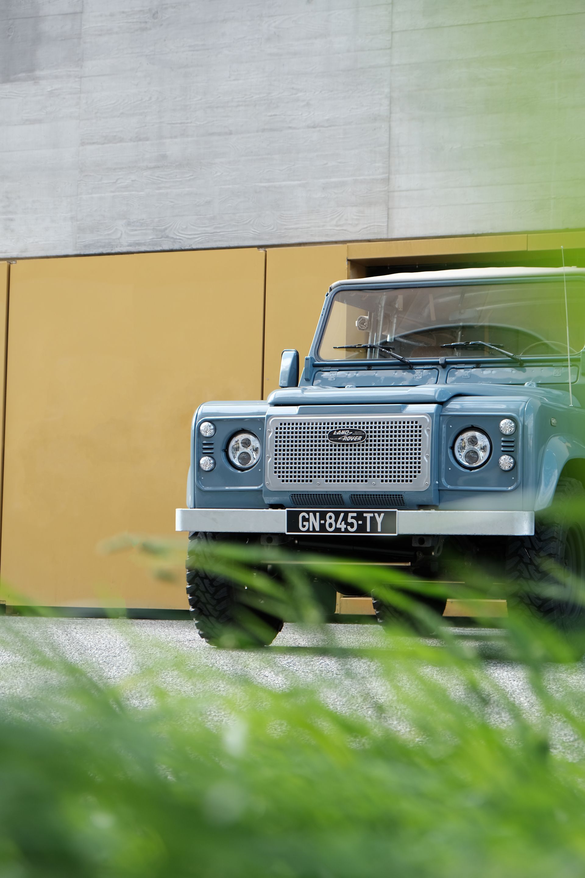 Land Rover Defender bleu garé devant un bâtiment jaune et gris, avec un feuillage vert au premier plan.