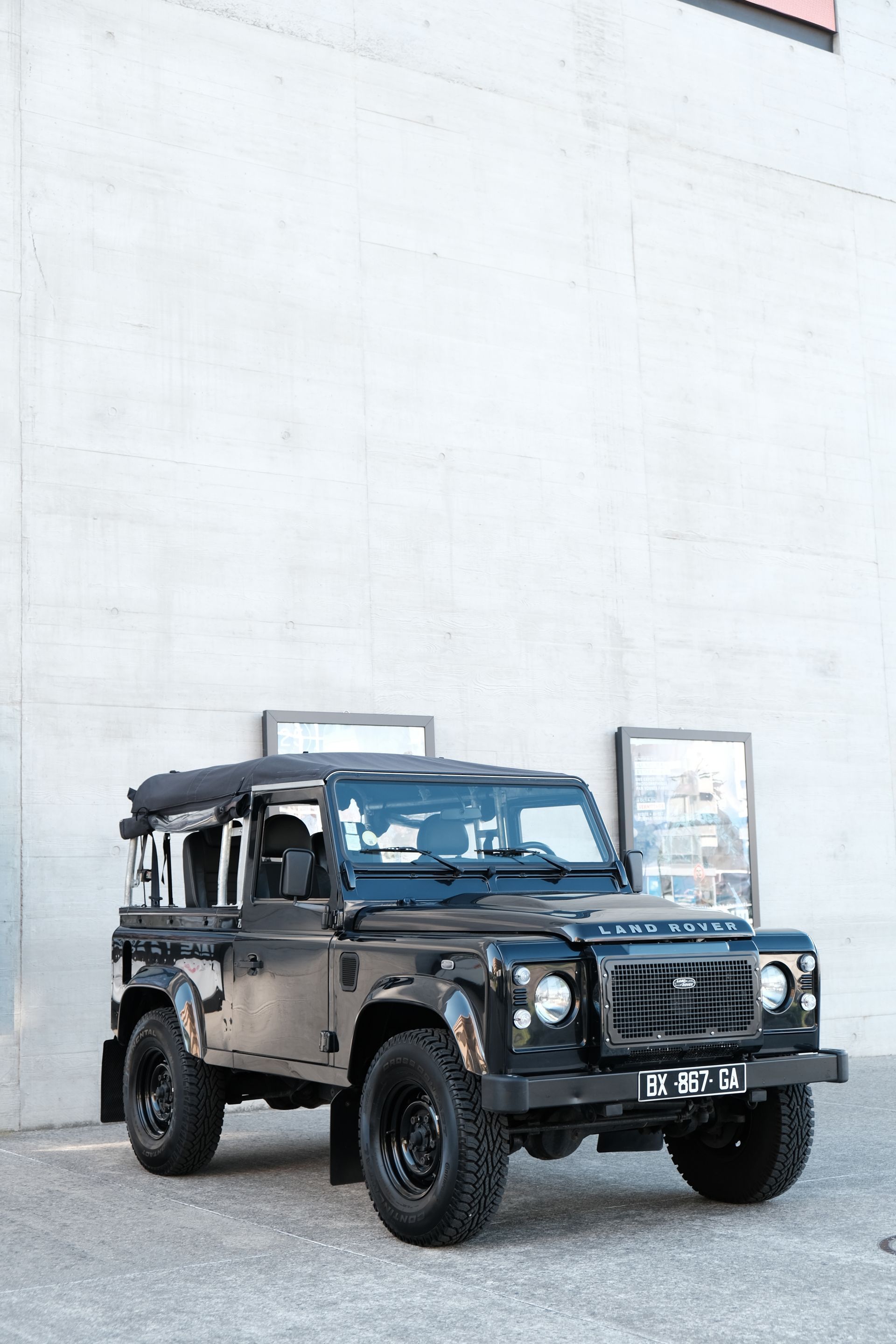 Land Rover Defender noir garé devant un mur en béton, avec deux affiches visibles.