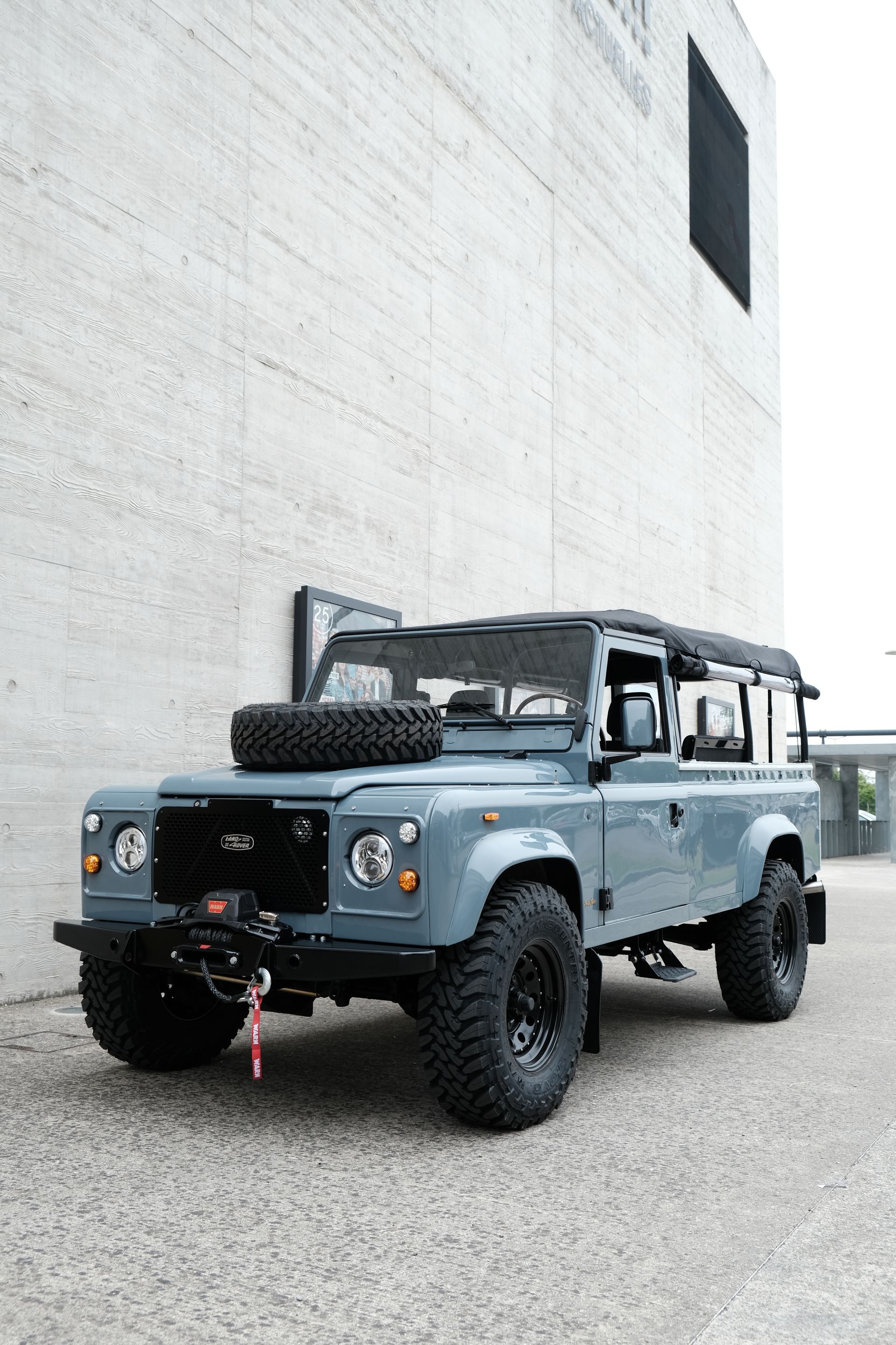 Land Rover Defender bleu garé devant un bâtiment en béton ; roue de secours sur le capot.