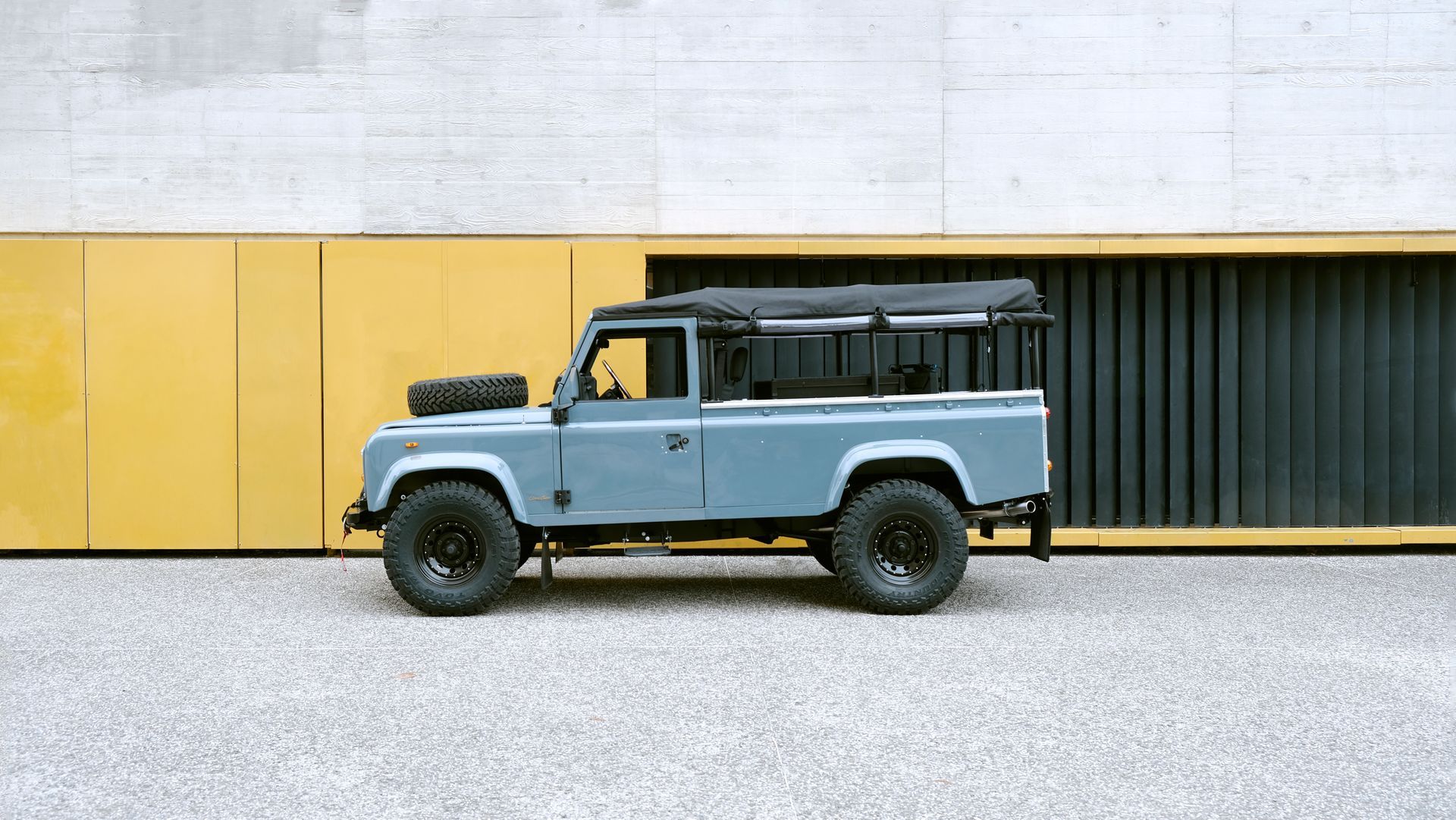 Land Rover Defender bleu garé contre un mur jaune et gris, avec une roue de secours sur le capot et une galerie de toit noire.
