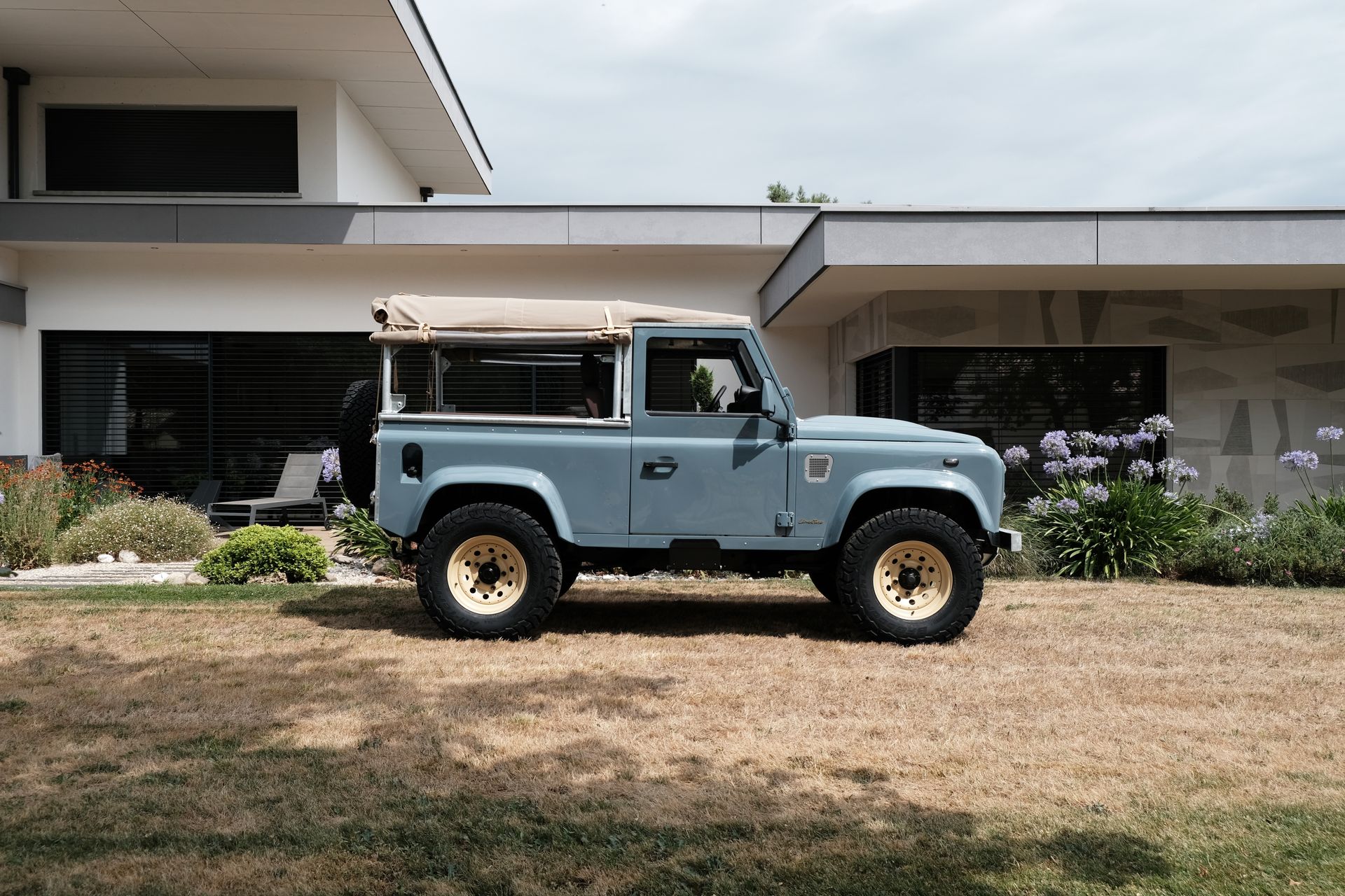 Land Rover Defender bleu clair avec toit beige, garé sur de l'herbe brune devant une maison moderne.