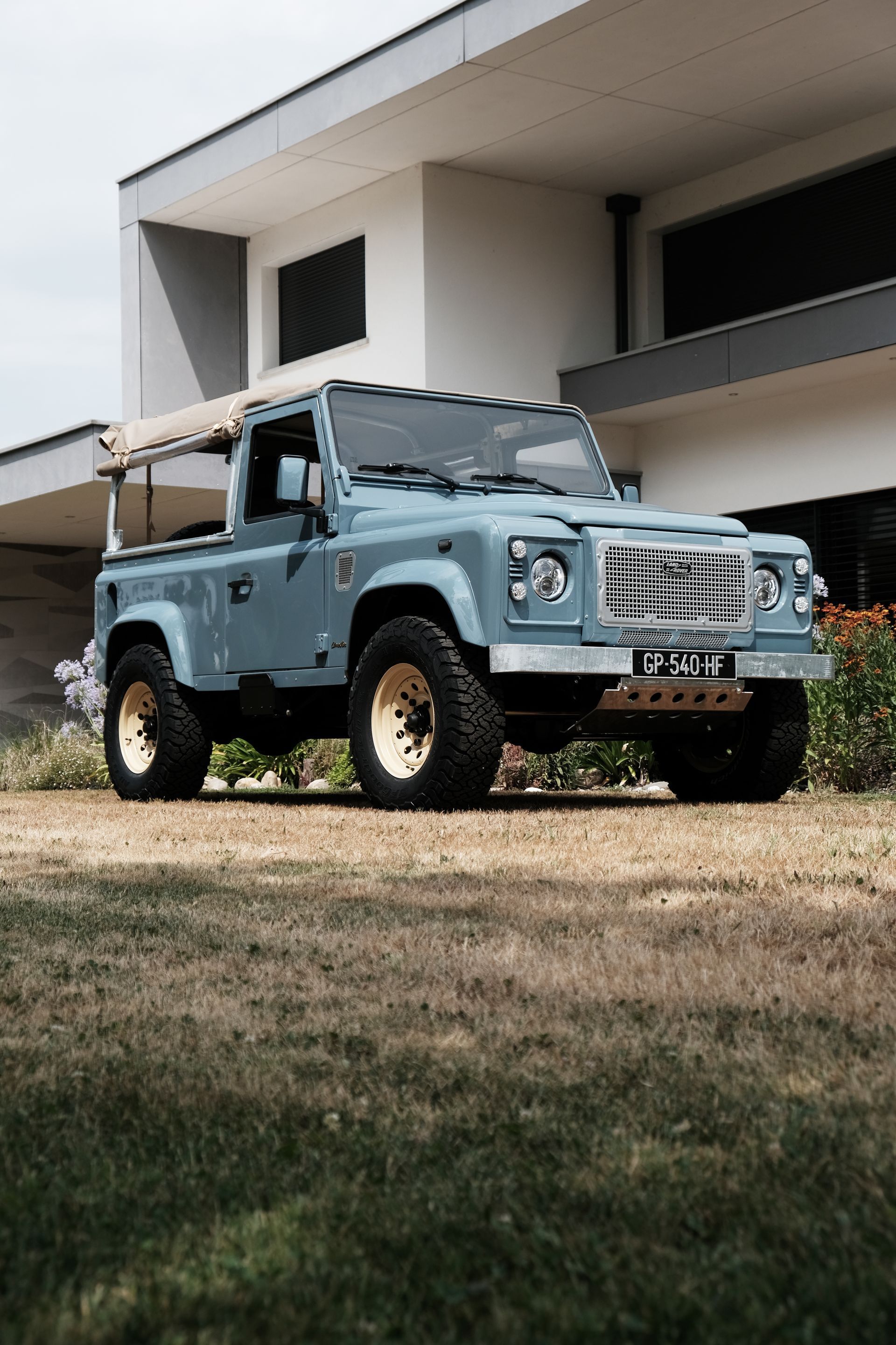 Land Rover Defender bleu garé sur l'herbe devant un bâtiment moderne avec des roues beige.