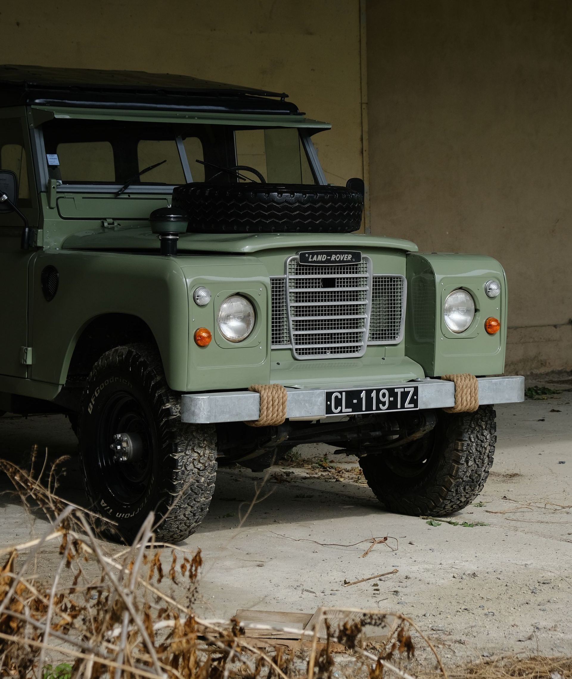 Land Rover vert vintage garé près d'un mur. Pneus noirs, roue de secours sur le capot et plaque d'immatriculation française.