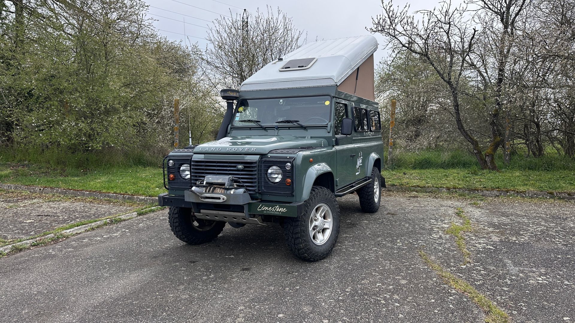 Land Rover Defender vert avec tente sur le toit sur une route pavée, devant des arbres.
