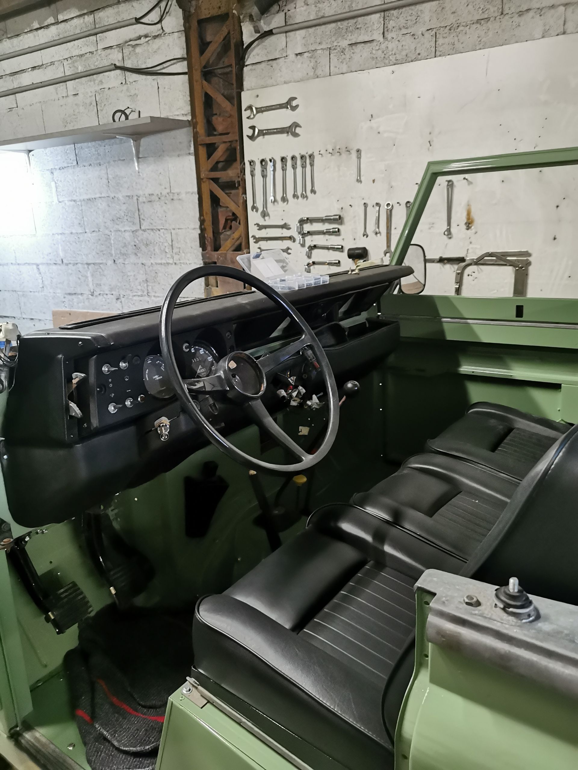 Intérieur d'un véhicule vintage vert, sièges noirs, volant, tableau de bord, outils au mur.