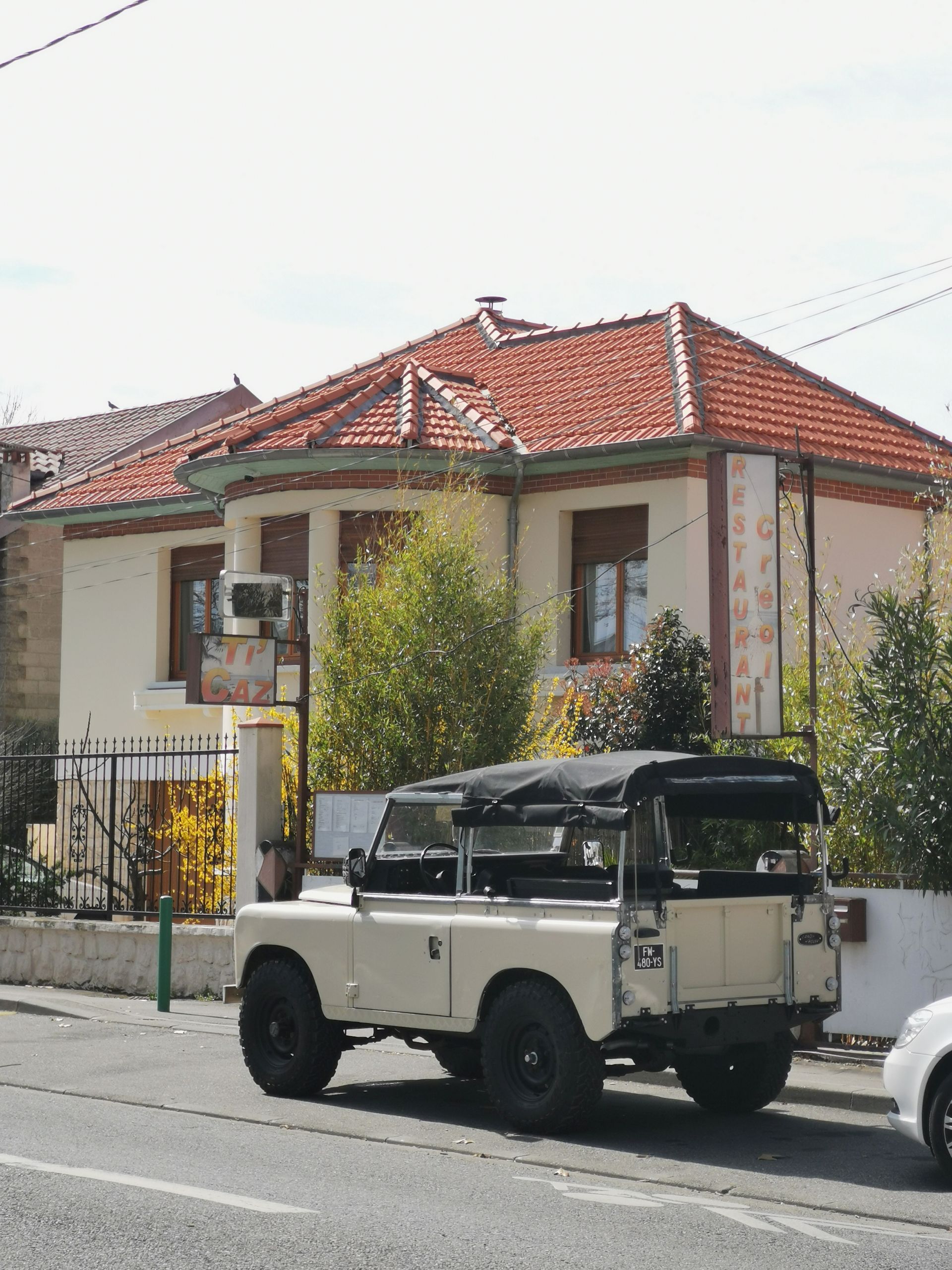 Land Rover beige garé devant un bâtiment de couleur claire avec un toit de tuiles rouges par une journée ensoleillée.