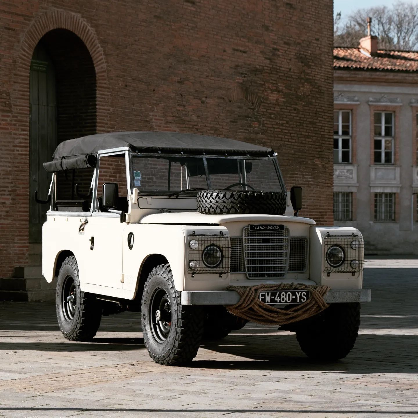 Crème Land Rover Defender garé sur une place avec un bâtiment en briques.
