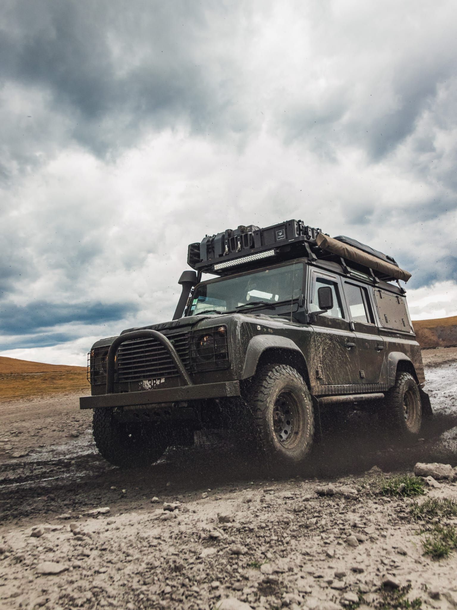 Un Land Rover Defender sombre et modifié roulant sur un terrain boueux sous un ciel nuageux.