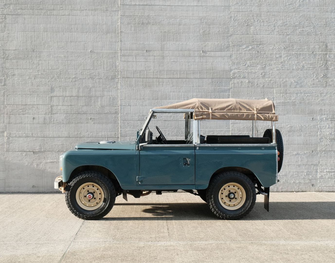Land Rover bleu vintage avec toit en toile beige garé devant un mur en béton.