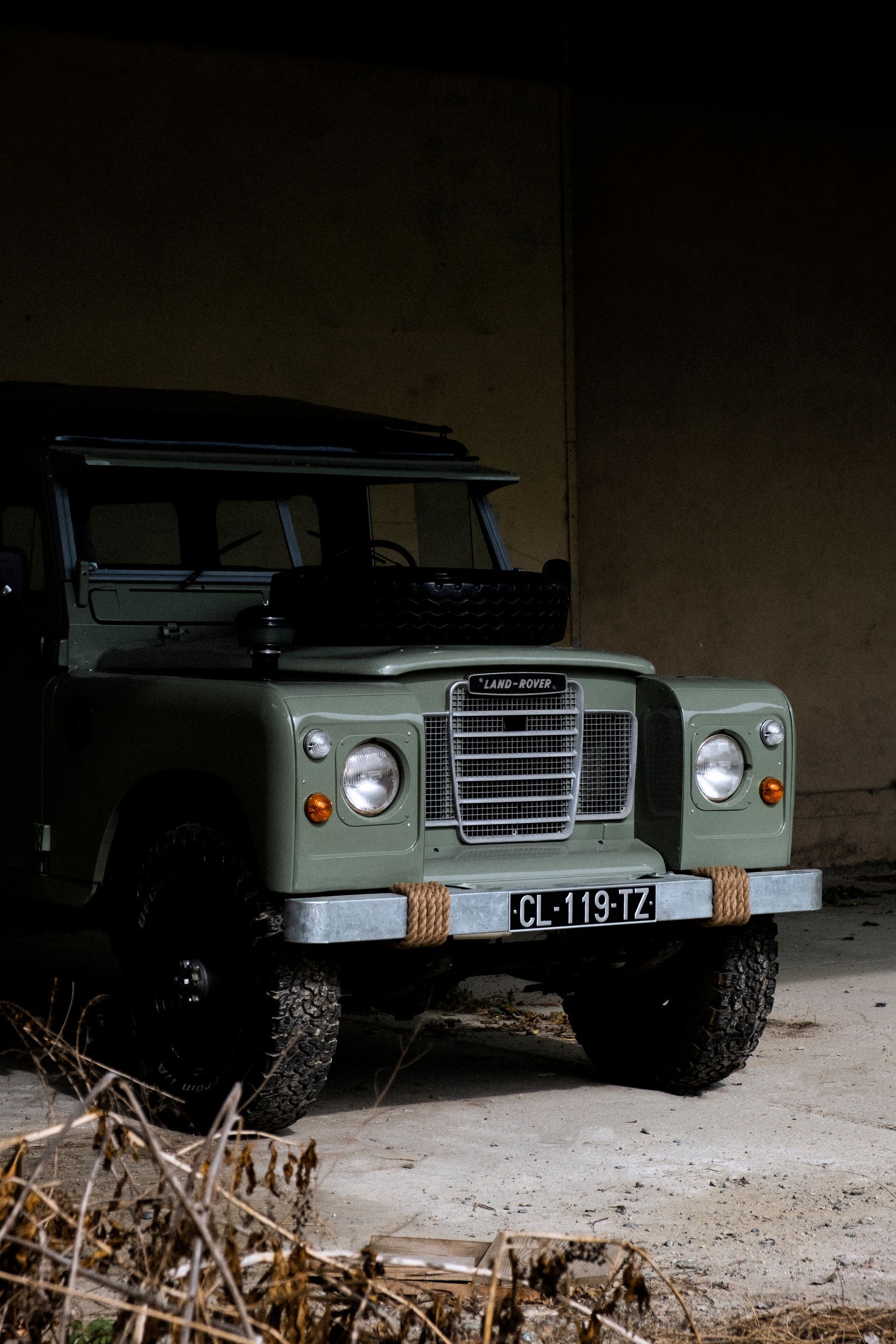Land Rover vert garé dans un hangar sombre ; pare-chocs avant rouillé ; pneus noirs.