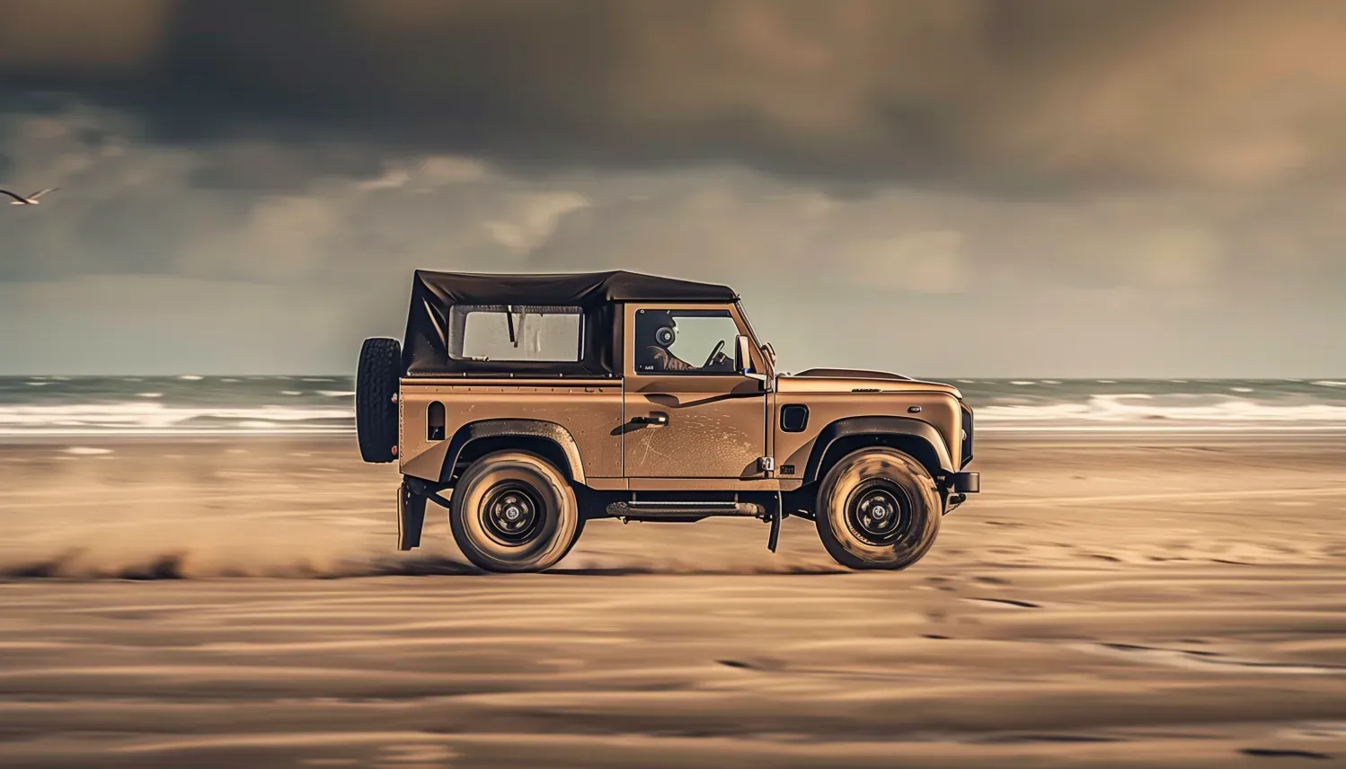 Un Land Rover Defender beige circule sur une plage de sable, avec du sable soulevé. Ciel sombre et océan en arrière-plan.