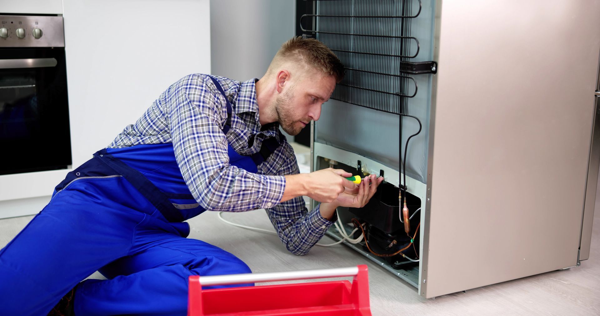 Technician in blue overalls repairing kitchen refrigerator with tools, kneeling near appliance.