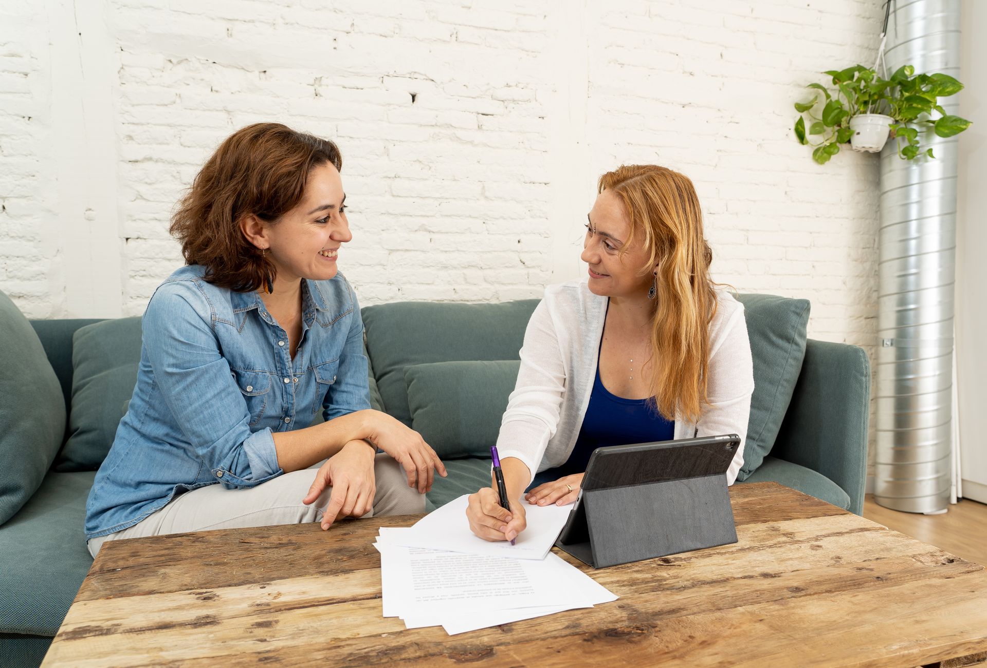 Two women sit at a table, discussing paperwork, with a tablet nearby in a bright living room setting