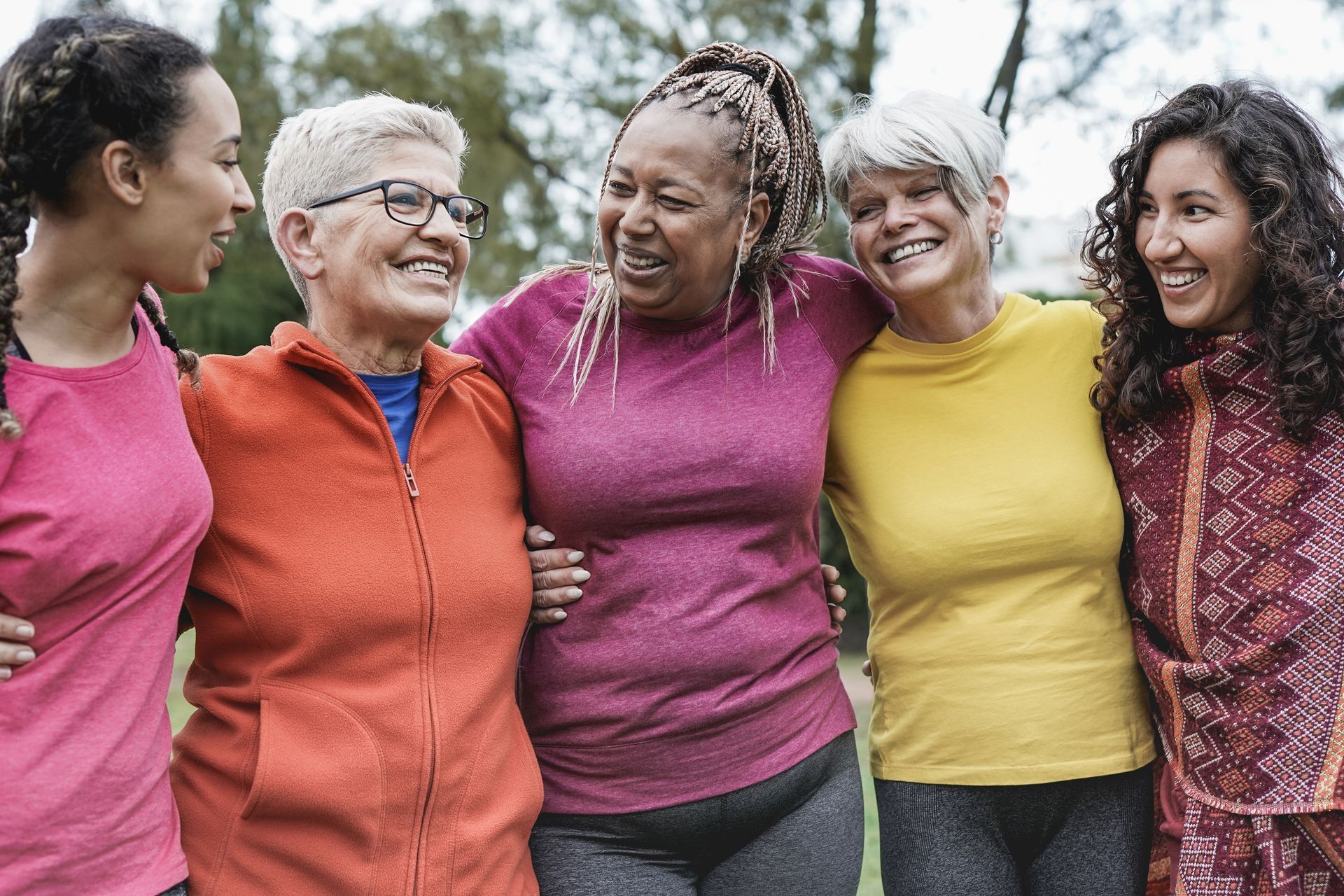 Five women smiling with arms around each other outdoors.