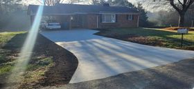 A freshly paved driveway leads to a brick house with a carport. The sky is bright.