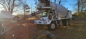 A white cement truck pouring concrete in a dirt area surrounded by trees under a blue sky.
