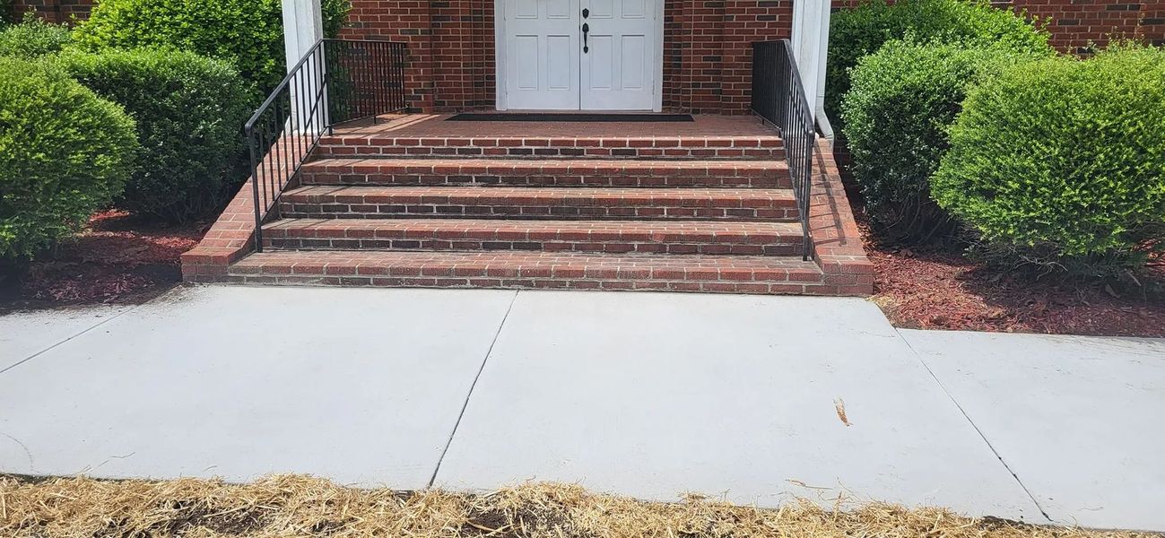 Brick steps leading to a white door, flanked by green bushes and black railings. A concrete sidewalk in front.