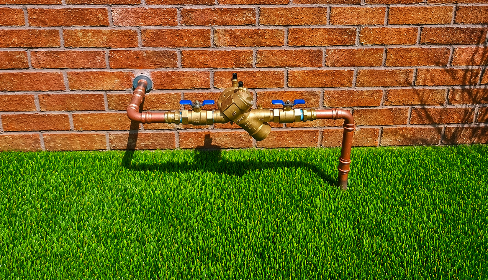 A brass backflow preventer valve with a blue cap, mounted on two metal pipes in a landscaped garden bed with mulch.
