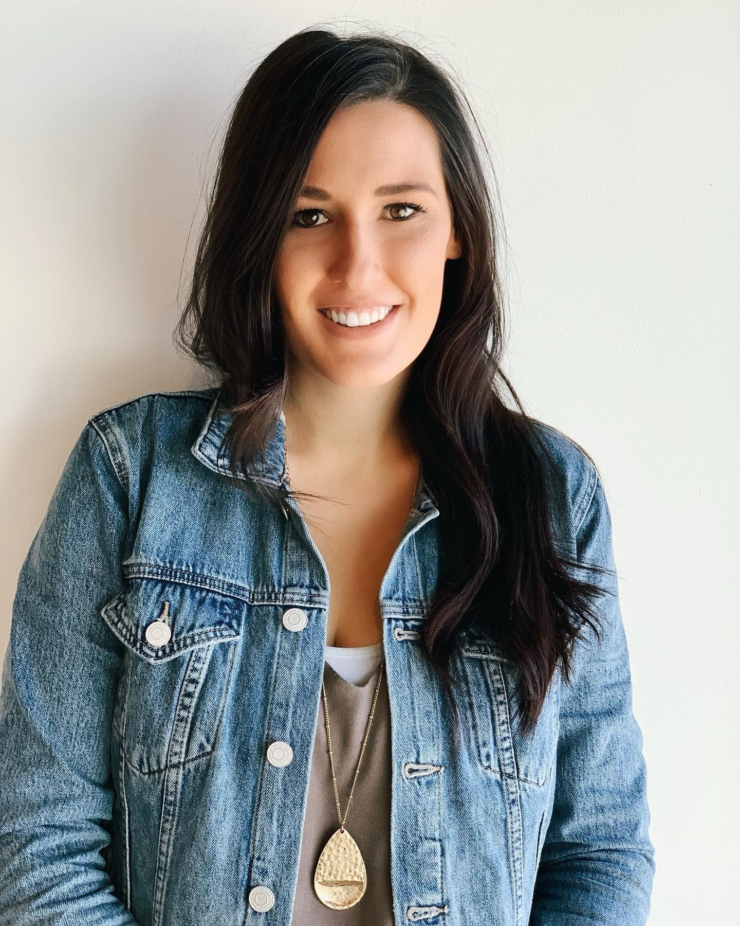 A woman wearing a denim jacket and a necklace smiles for the camera.