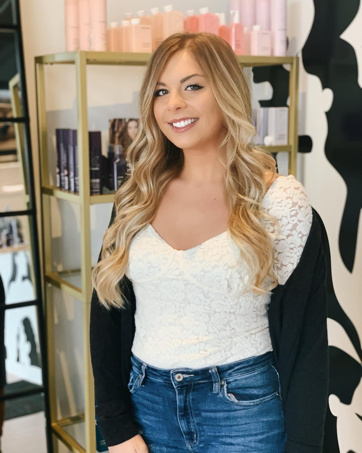 A woman in a white top and blue jeans is standing in front of a shelf.