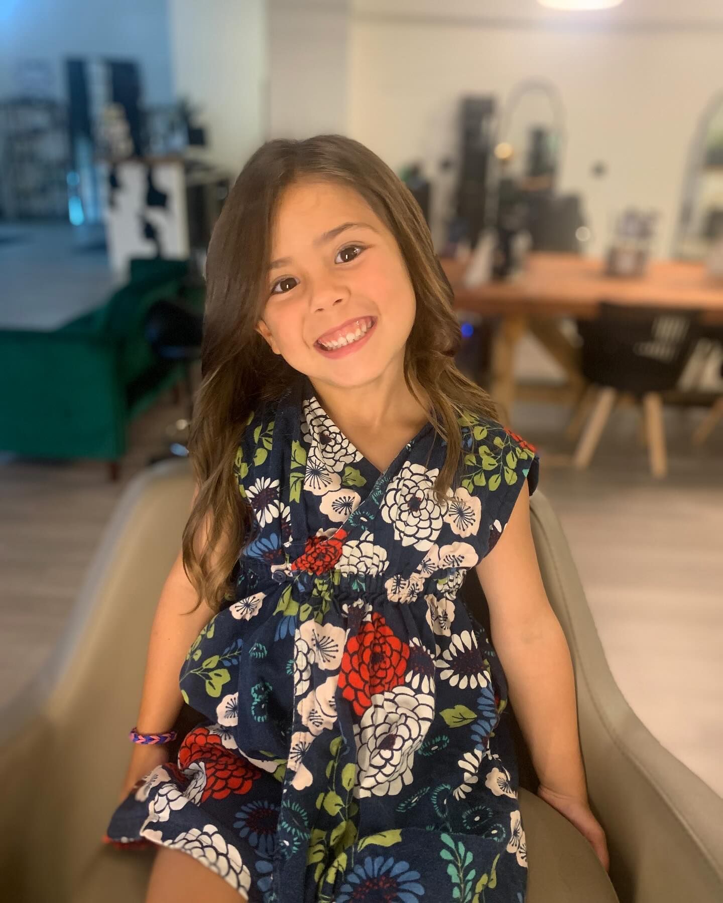 A little girl in a floral dress is sitting in a chair and smiling.