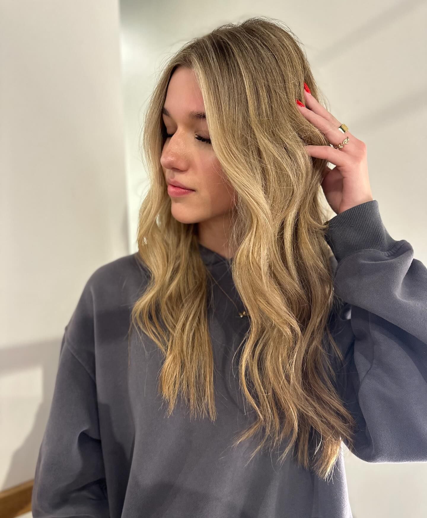 A woman with long blonde hair is wearing a gray sweatshirt and touching her hair.