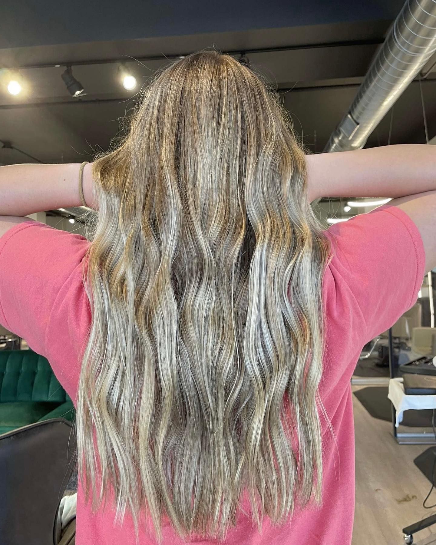 A woman with long blonde hair is standing in a salon with her hands behind her head.