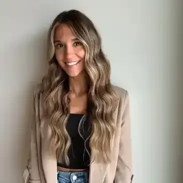 A woman with long hair is wearing a tan jacket and smiling.