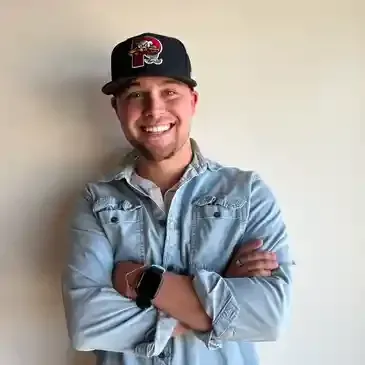 A man wearing a hat and a denim shirt is standing with his arms crossed and smiling.
