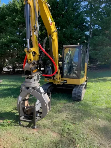 Yellow excavator with grapple attachment on grass, trees in background.