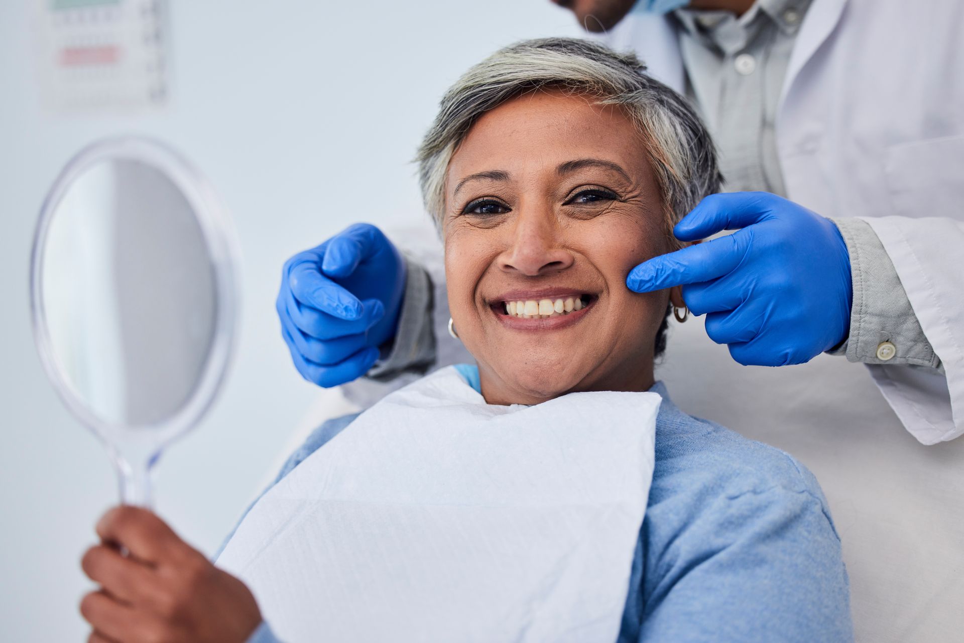 Smiling patient in dental chair with mirror while dentist adjusts blue-gloved hands nearby