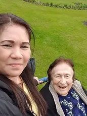 Woman takes a selfie with an elderly woman seated outdoors on green grass. Both smiling.