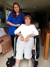 Woman in a wheelchair with another woman standing behind her; both are smiling in a home setting.
