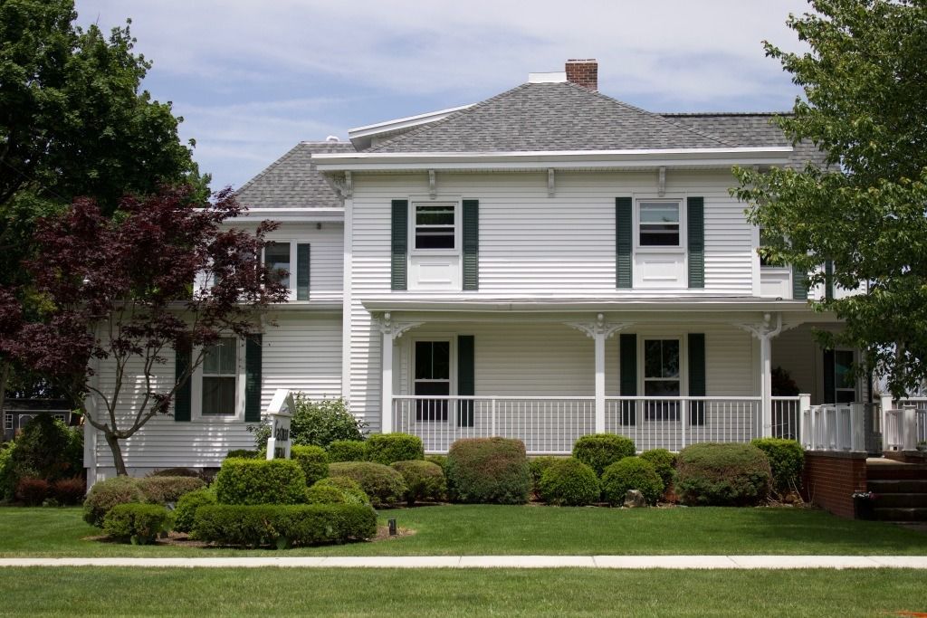 White, two-story house with green shutters, front porch, and trimmed bushes on a green lawn.