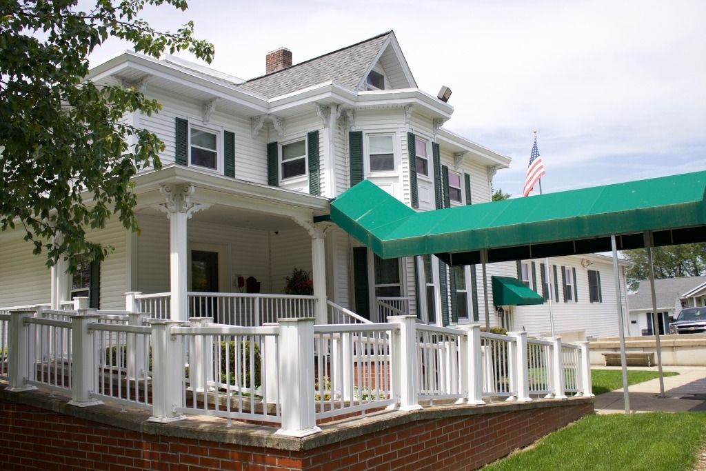 White two-story building with green awning, porch, and American flag on a sunny day.