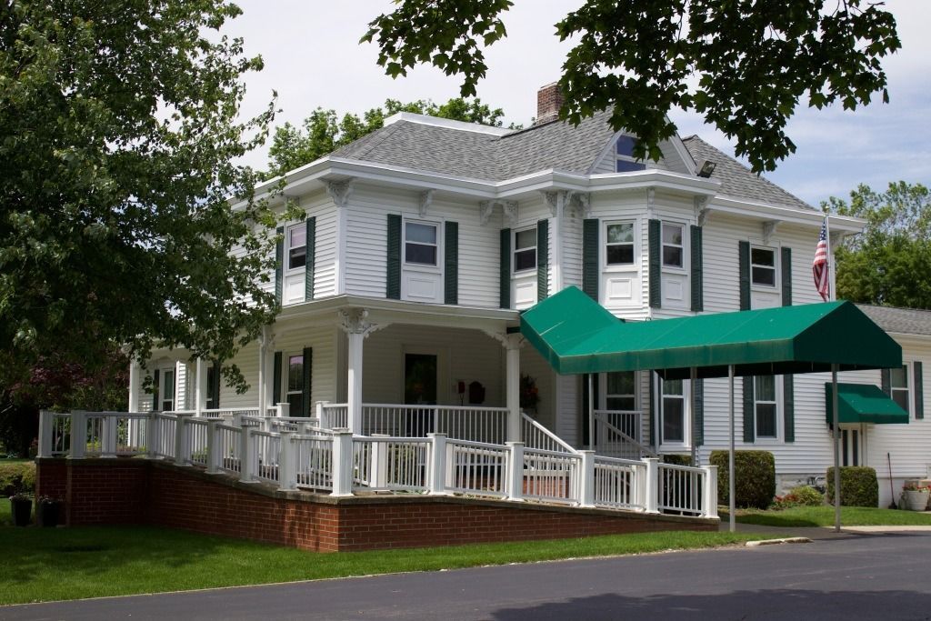 White two-story building with green shutters and awning. A ramp and porch are in front.