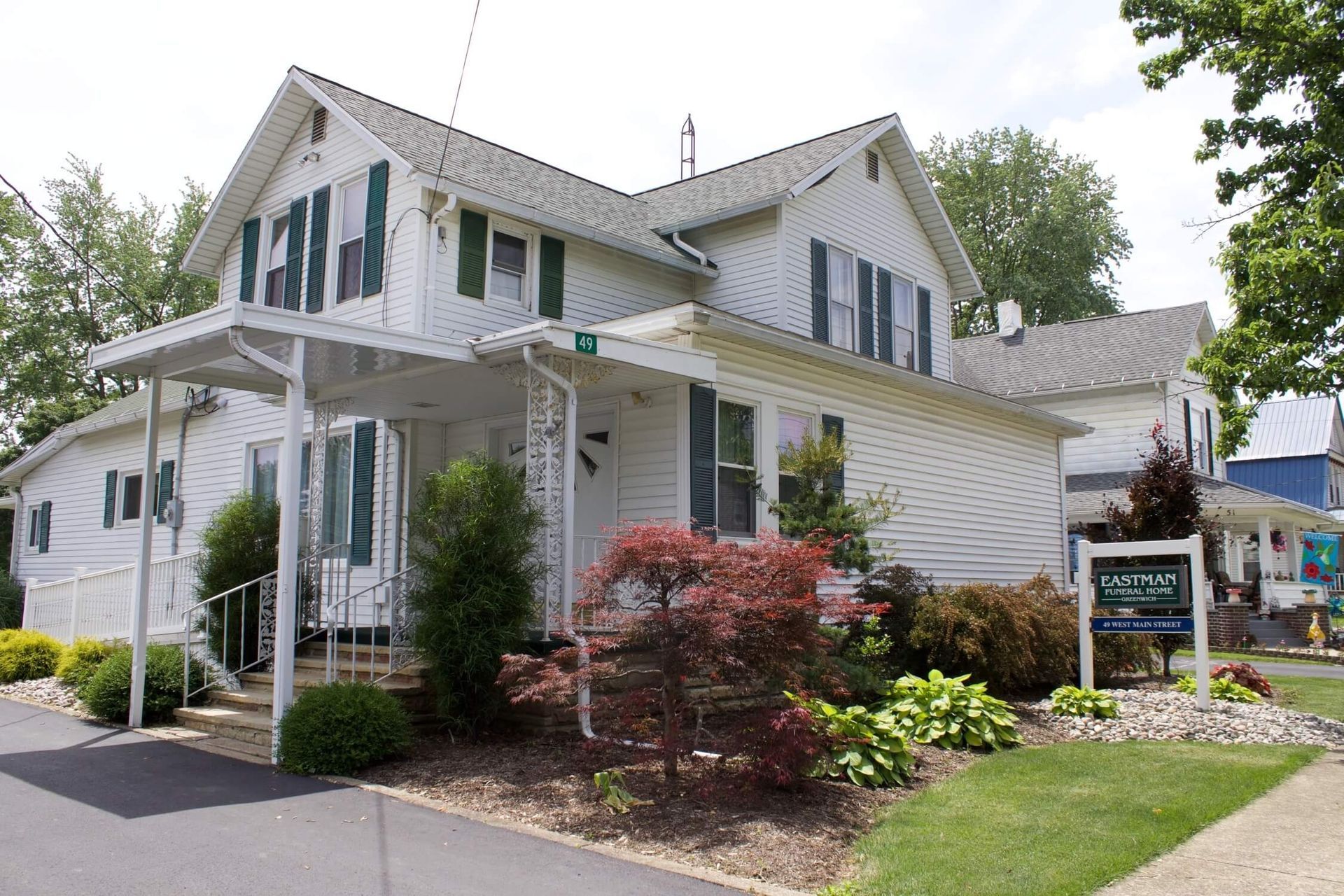White two-story house with green shutters, a covered entrance, and a sign in the front yard.